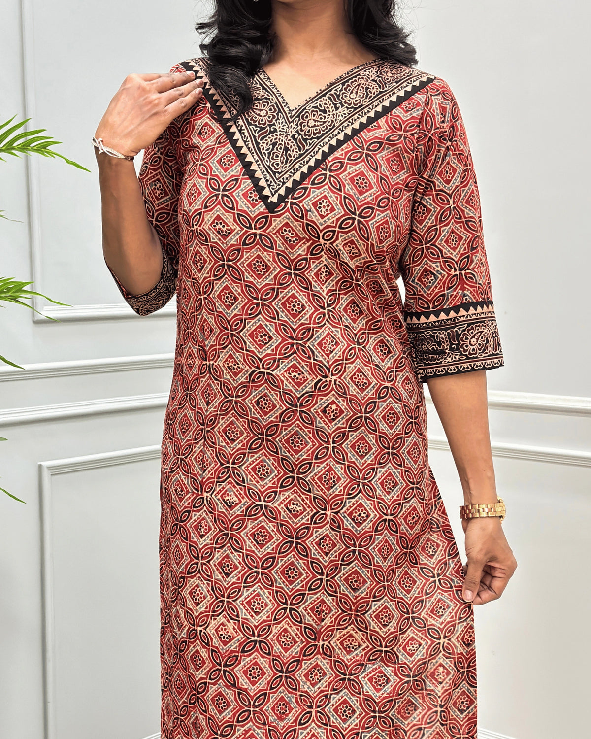 Close-up of brown and red Hand-block Ajrakh pure cotton Long-kurta with a Ajrakh geometric pattern, featuring a V-neckline and patterned sleeve cuffs, standing against a white paneled wall