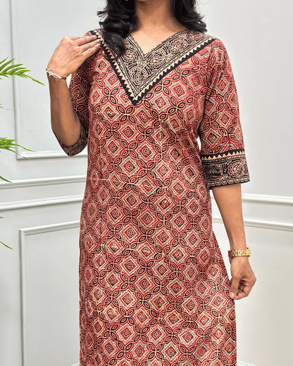 Close-up of brown and red Hand-block Ajrakh pure cotton Long-kurta with a Ajrakh geometric pattern, featuring a V-neckline and patterned sleeve cuffs, standing against a white paneled wall