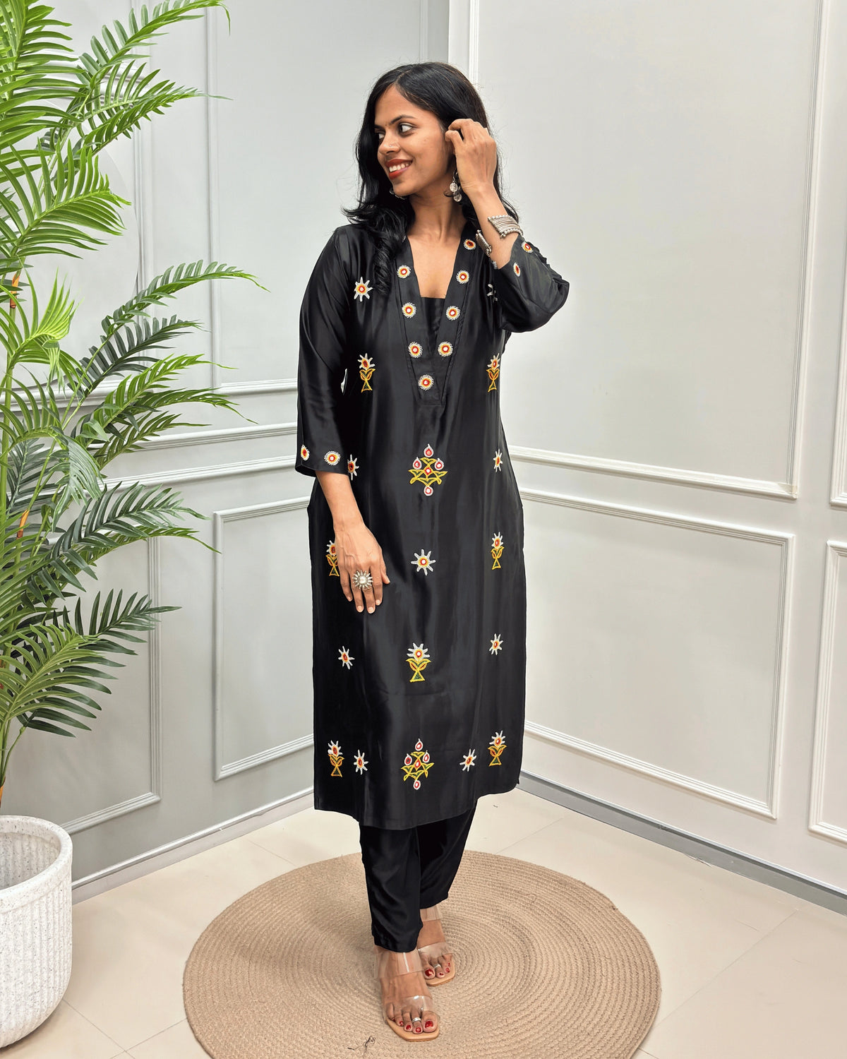 Black women's Pure Mashru Silk Long Kurti with yellow and white floral embroidery, & Ahir work on front & Sleeves cuffs , paired with Black pants, standing on a round woven mat in front of a white paneled wall with a green plant to the side.