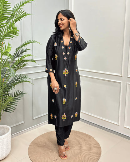 Black women's Pure Mashru Silk Long Kurti with yellow and white floral embroidery, & Ahir work on front & Sleeves cuffs , paired with Black pants, standing on a round woven mat in front of a white paneled wall with a green plant to the side.