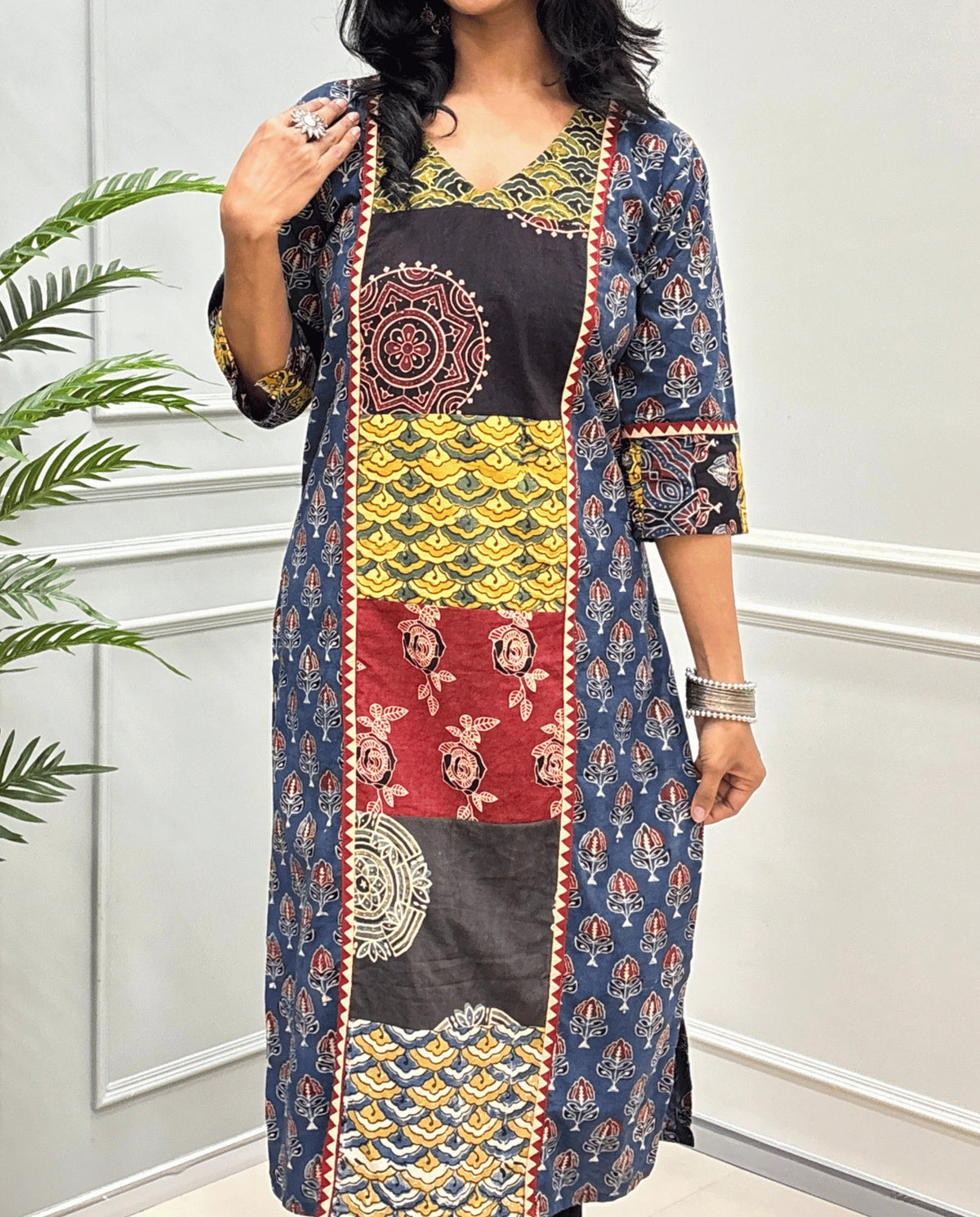 Close-up of Blue Women pure cotton long kurti featured with blue, yellow, red, and black Geometric different print panels on Front & Sleeves cuffs , Ajrakh print on blue base outside the middle panel , three quarter Sleeves & V-neck standing indoors against a light paneled wall with a green plant on the left.