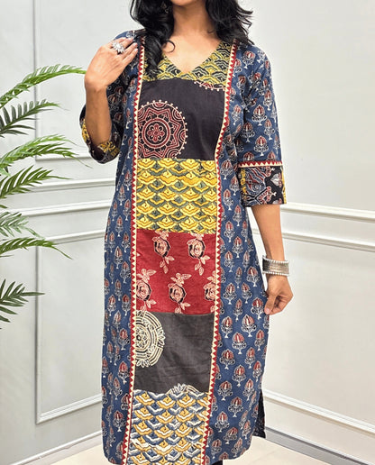 Close-up of Blue Women pure cotton long kurti featured with blue, yellow, red, and black Geometric different print panels on Front & Sleeves cuffs , Ajrakh print on blue base outside the middle panel , three quarter Sleeves & V-neck standing indoors against a light paneled wall with a green plant on the left.