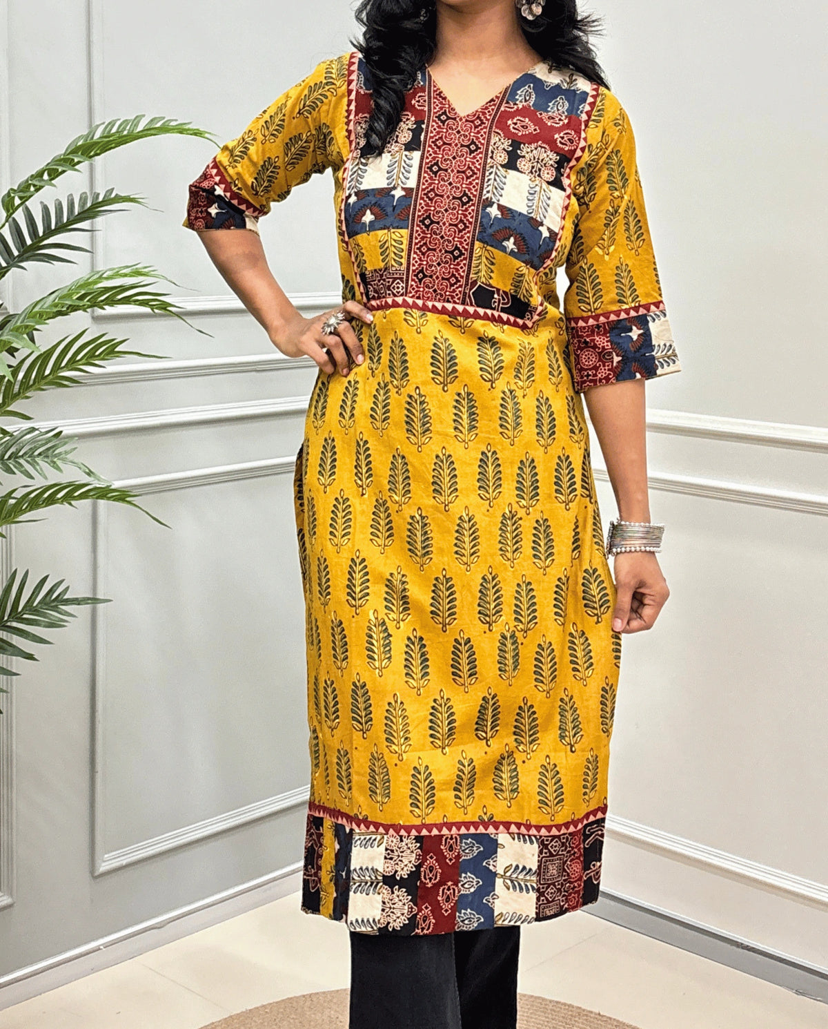 Close-up of Yellow Women's Hand-block Ajrakh Pure cotton Long-Kurti featured with 3/4th quarter Sleeves with multicolor patchwork-style yoke on Front, Cuffs and hem, V-shaped neck, Ajrakh print on front & Sleeves, paired with black Flared pants, standing indoors against a white paneled wall next to a potted plant.