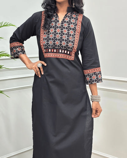 Close-up of Black Hand-block Ajrakh Pure cotton Long kurti with three quarter sleeves with printed cuffs,  intricate black & red geometric embroidery, with hand mirror work and thread work on border of mirror , with Mandarian collar V-neck and sleeve cuffs, standing against a light gray paneled wall.