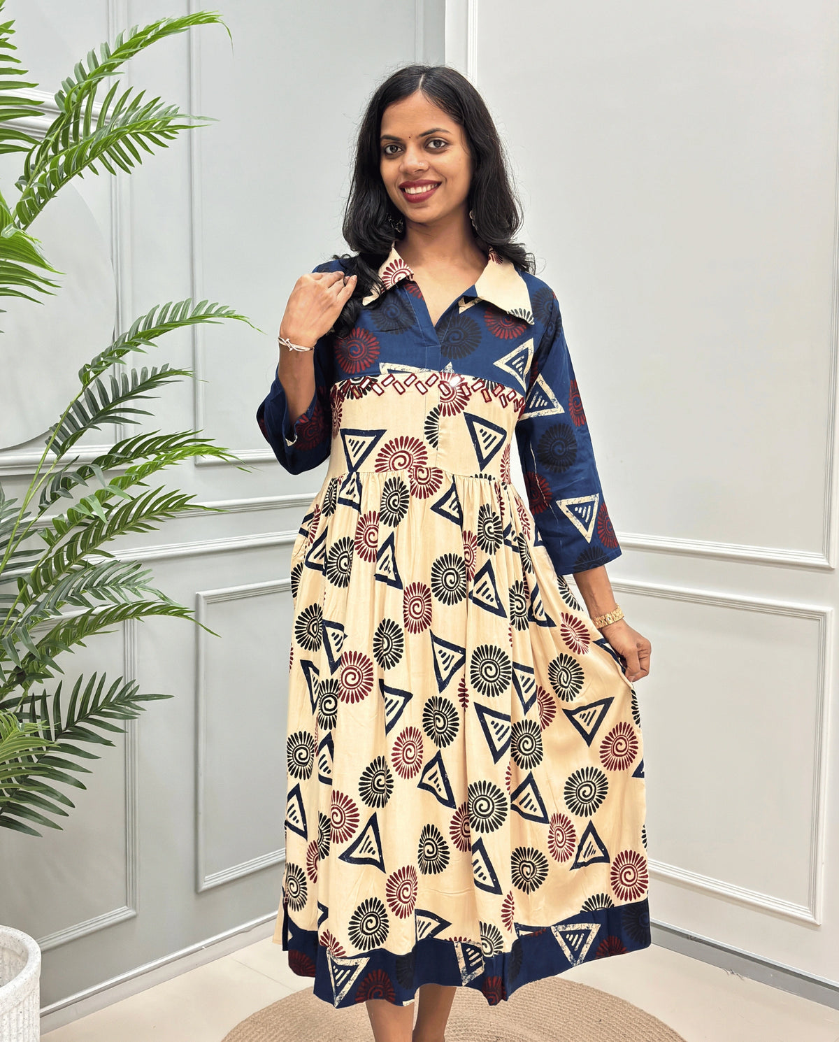 Beige Women's Ajrakh Vegetable dyed plant based Cotton Long-Kurti with a Blue neck and cuffs, and a Beige-colored collar & dress featuring a geometric pattern of triangles and spiral in Red, black, and blue, Hand-mirror work on front, standing on a round woven rug in front of a white paneled wall with a green potted plant on the left.