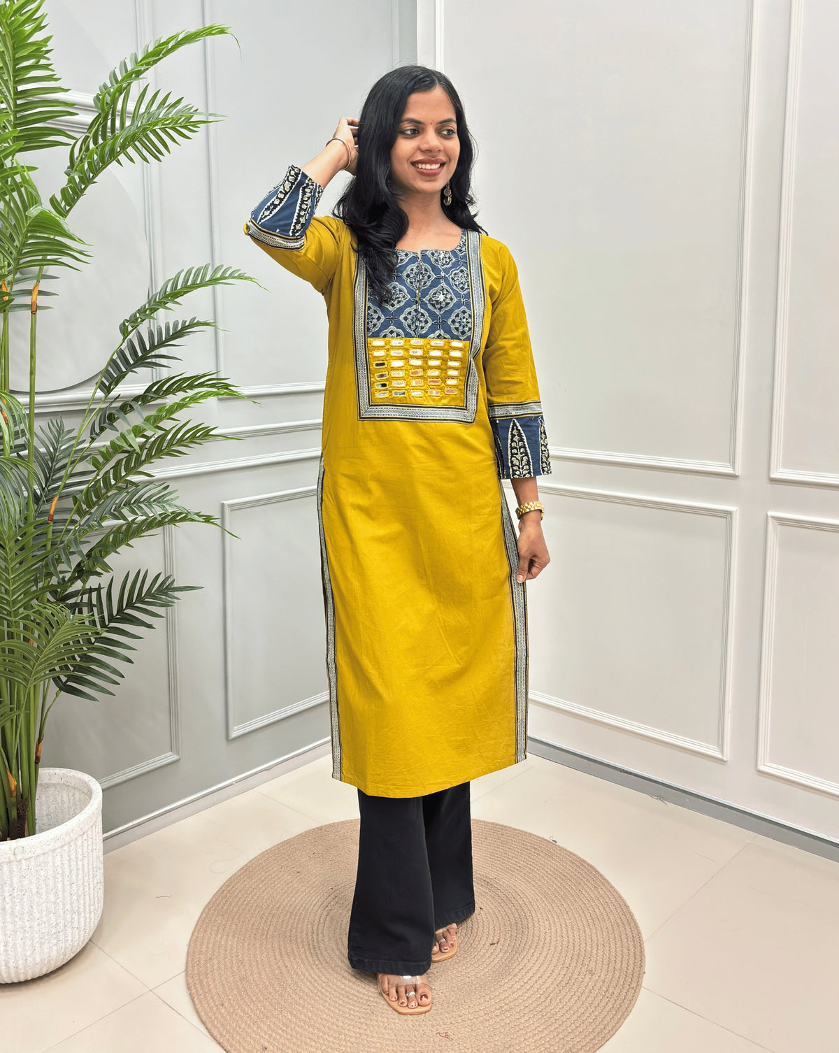 Yellow Women's Hand-block Ajrakh Pure Cotton Long Kurti with a red base and abstract Blue, Black & Beige  geometric embroidery on the chest and sleeve cuffs, paired with black Flared pants, standing in a decorated room with a potted plant.