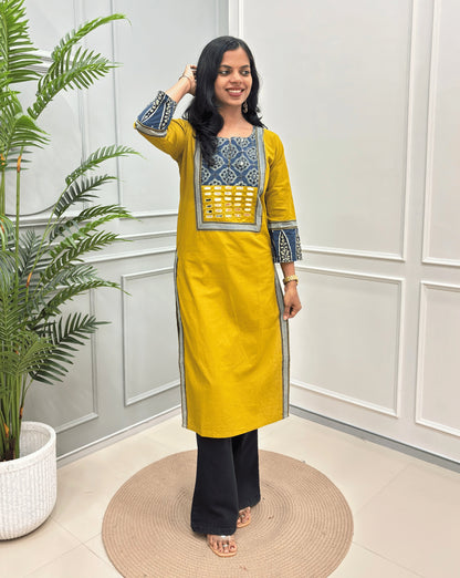 Yellow Women's Hand-block Ajrakh Pure Cotton Long Kurti with a red base and abstract Blue, Black & Beige  geometric embroidery on the chest and sleeve cuffs, paired with black Flared pants, standing in a decorated room with a potted plant.