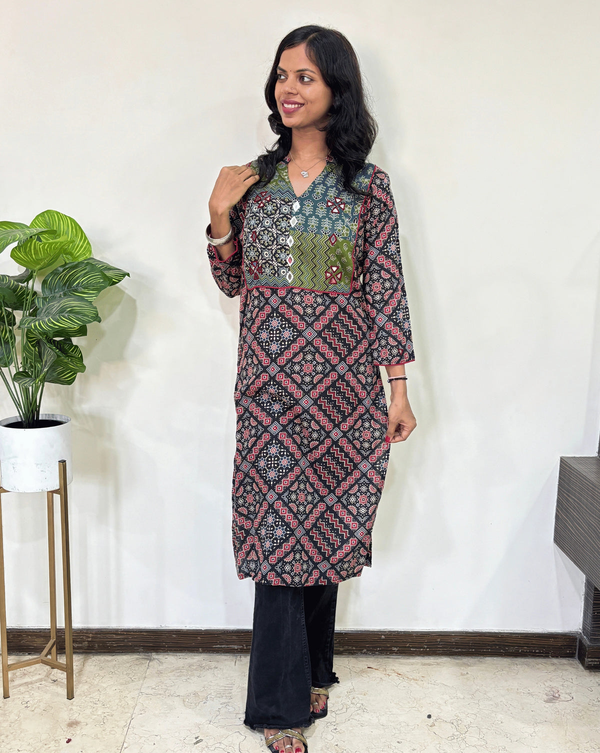 Black Women's Ajrakh Vegetable Dyed plant based cotton long kurti with green, black, and red patterns, paired with black flared pants, standing in a room with a white wall and a potted plant.