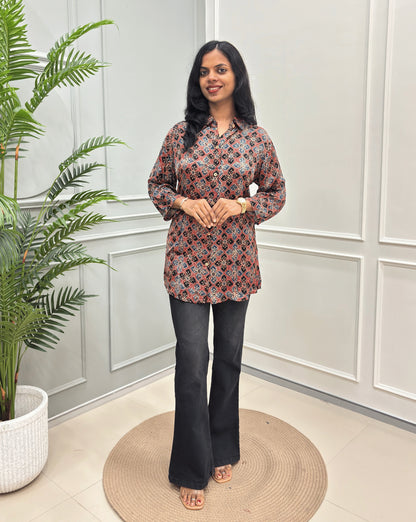 Red Women's Ajrakh Pure Modal Silk Shirt with a Floral print , black flared pants, standing on a round woven rug in front of a white paneled wall with a potted plant on the left.