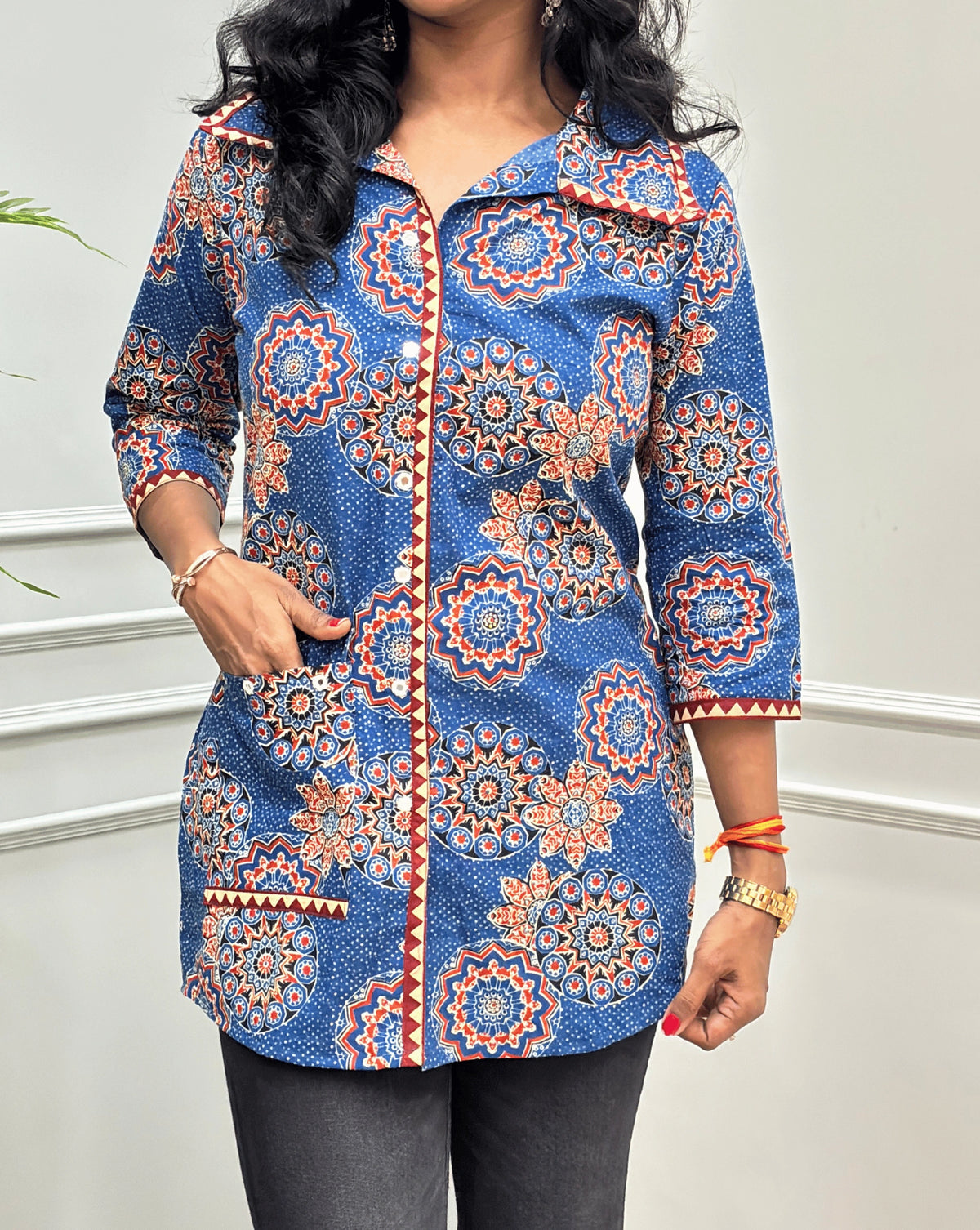 Close-up of Blue women's Ajrakh pure cotton Shirt featured with Three Quarter Sleeves & Collar V shaped neck  with a Blue base and Multi color floral print, Hand-mirrorwork on front button style, Sleeves cuffs & pocket, paired with black flared pants. Standing on a round woven rug against a light paneled wall with a potted plant at left.