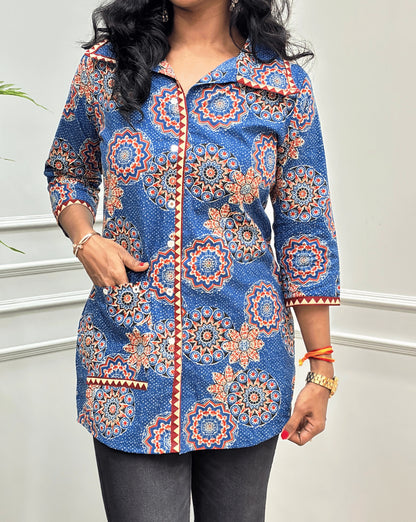 Close-up of Blue women's Ajrakh pure cotton Shirt featured with Three Quarter Sleeves & Collar V shaped neck  with a Blue base and Multi color floral print, Hand-mirrorwork on front button style, Sleeves cuffs & pocket, paired with black flared pants. Standing on a round woven rug against a light paneled wall with a potted plant at left.