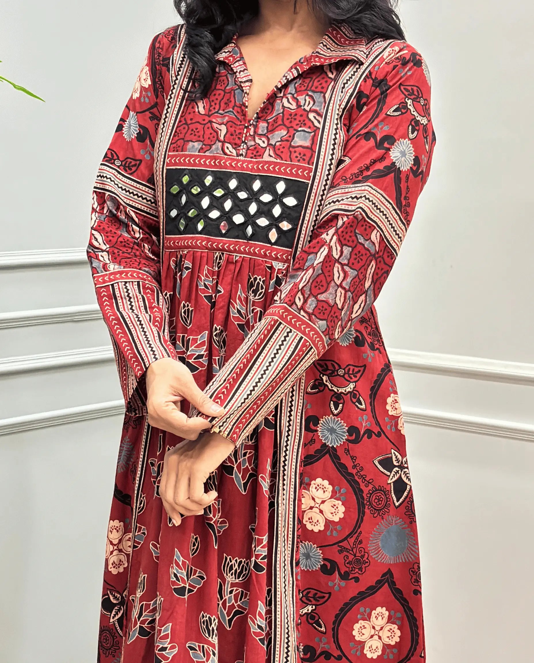 Close-up of handcrafted mirror work on red Ajrakh cotton kurti along with Ajrakh printed Sleeves detail on long kurti , standing against a white paneled wall.