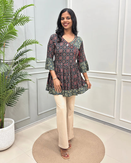 Black Women's Ajrakh Vegetable Dyed plant based cotton Short Kurti with a teal and red geometric pattern, paired with beige flared pants, standing on a round woven mat in front of a white paneled wall with a potted plant.