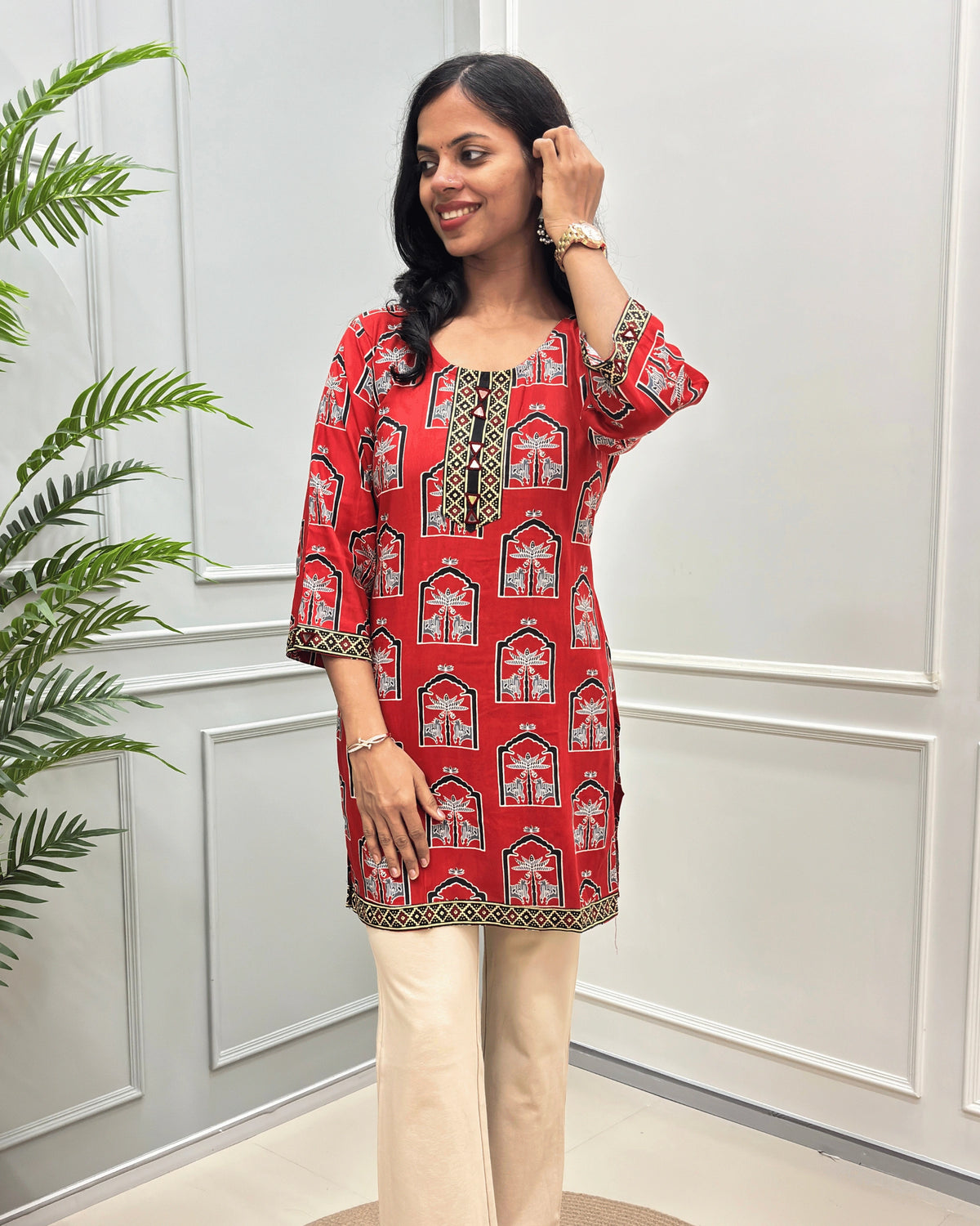 Front view of Red Women's Ajrakh plant based Cotton Short Kurti with a red and Grey Pichhwai pattern, paired with beige flared pant , standing in front of a white paneled wall with a green plant on the left.