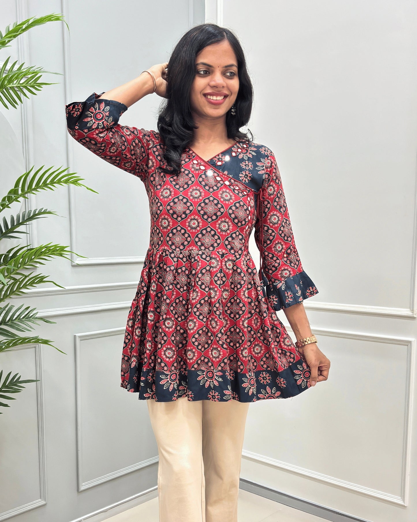 Front view of Red Ajrakh plant based rayon cotton Short Kurti featured with Ajrakh  geometric patterned with Navy-blue borders on neck , cuffs & bottom  paired with beige pants, standing in front of a white paneled wall with a green plant on the left.