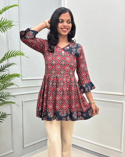 Front view of Red Ajrakh plant based rayon cotton Short Kurti featured with Ajrakh  geometric patterned with Navy-blue borders on neck , cuffs & bottom  paired with beige pants, standing in front of a white paneled wall with a green plant on the left.