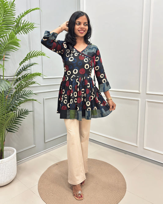 Black Women's Ajrakh Vegetable dyed plant based Cotton Short-Kurti with three-quarter frill sleeves, and waist to bottom,  paired with beige flared pant , standing on a round woven rug in front of a white paneled wall with a potted plant on the left.