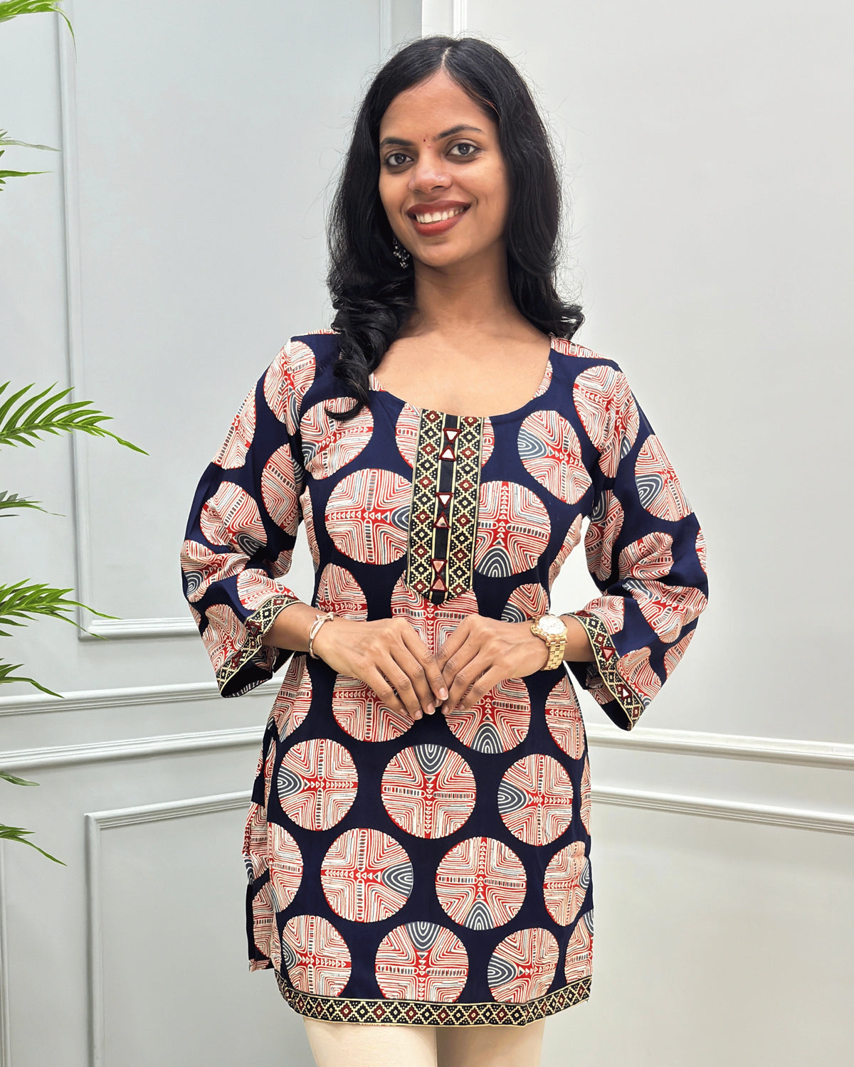 Close-up of Navy-blue plant based cotton short kurti with a red, beige, and white circular geometric pattern, Hand-mirror work on front and sleeves. standing against a light gray paneled wall. 