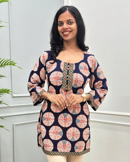 Close-up of Navy-blue plant based cotton short kurti with a red, beige, and white circular geometric pattern, Hand-mirror work on front and sleeves. standing against a light gray paneled wall. 