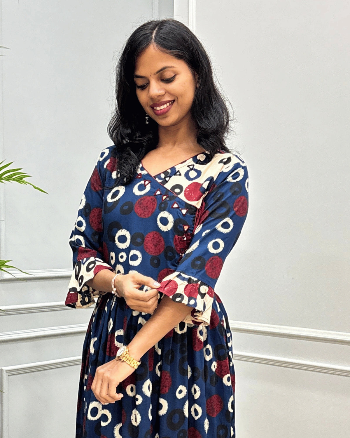 Close-up of Blue women's plant based cotton long kurti featured with red, beige and black circular print on blue base, hand-mirror work on round neck  standing in front of a light gray paneled wall.