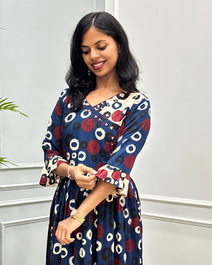Close-up of Blue women's plant based cotton long kurti featured with red, beige and black circular print on blue base, hand-mirror work on round neck  standing in front of a light gray paneled wall.