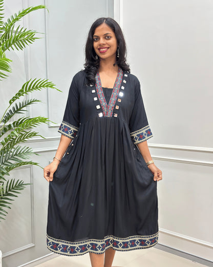 Front view of Black women's plant based cotton long-kurti with colorful embroidered details on the neckline, cuffs, and hem, standing in front of a white paneled wall with a green plant on the left.