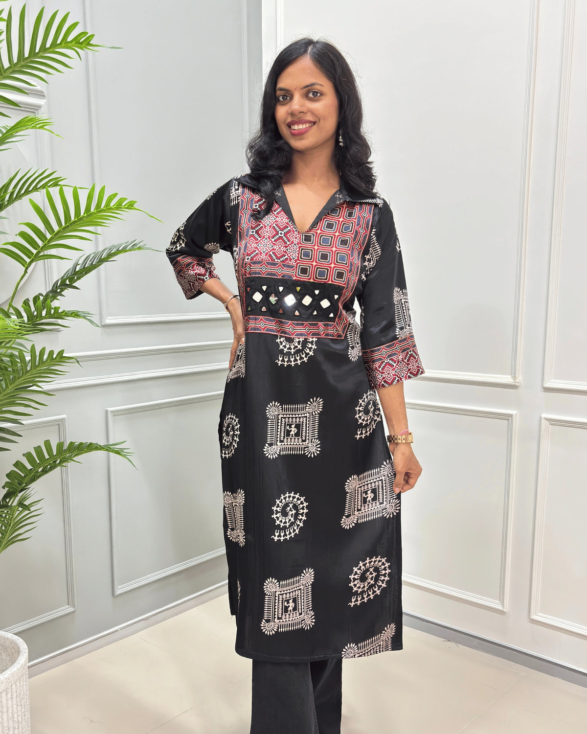 Front view of Black Ajrakh pure Mashru Silk Long kurti with geometric print in red, white, and beige on Front & sleeves, standing in a bright room with a white paneled wall and a potted plant.