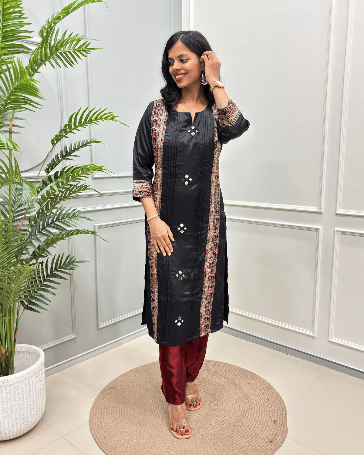 Black Women's Ajrakh Mashru Silk Long-kurti with patterned borders and Hand-mirror embellishments, paired with maroon pants, standing in a bright room with a potted plant and paneled wall.