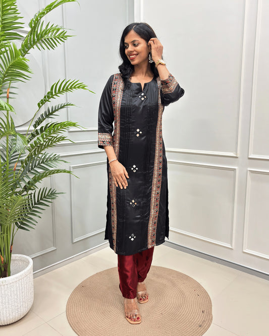 Black Women's Ajrakh Mashru Silk Long-kurti with patterned borders and Hand-mirror embellishments, paired with maroon pants, standing in a bright room with a potted plant and paneled wall.