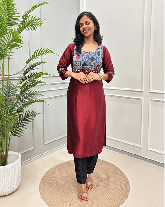 Red Women's Ajrakh Mahru Silk Long-kurti with a geometric Print yoke in blue, red, and white, with Hand-Mirror Work,  paired with black pants, standing on a round woven mat in front of a white paneled wall with a potted plant.