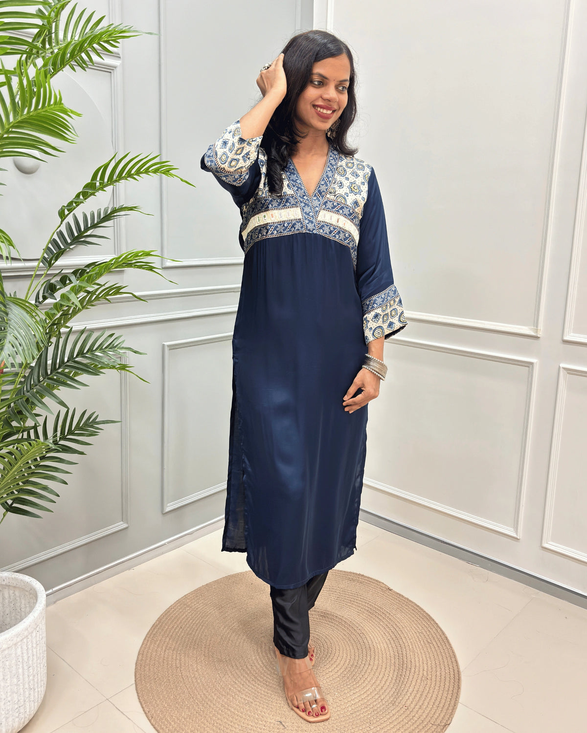 Navy Blue Women's Ajrakh Modal Silk Long-Kurti with intricate white base & Ajrakh print on the yoke and cuffs, paired with black pants and clear heeled sandals, standing on a round woven mat in a decorated room.