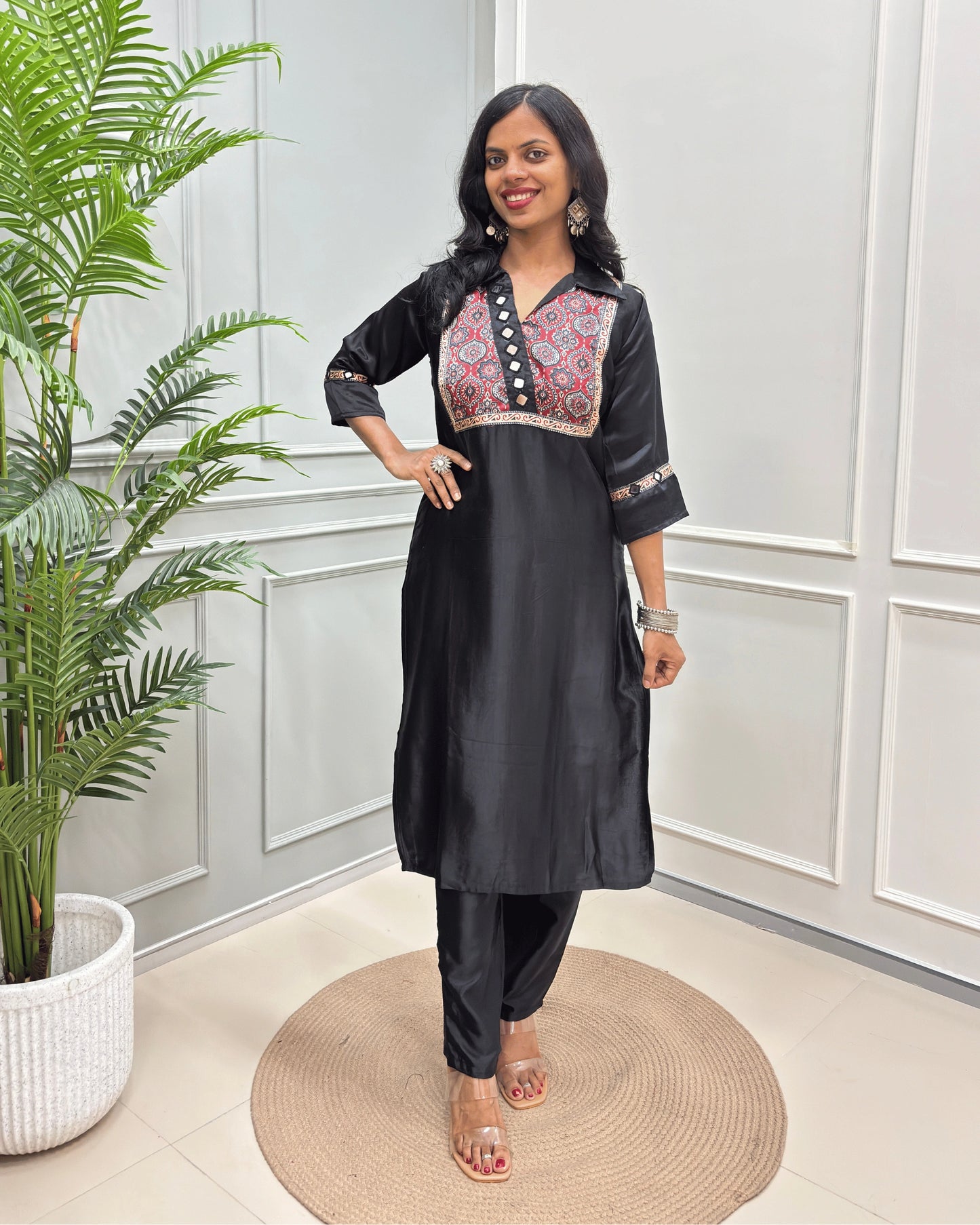 Black Women's Ajrakh Mashru Silk Long Kurti with colorful embroidered detailing on the chest and sleeves, paired with black pants, standing in a decorated room with a potted plant and paneled wall. 