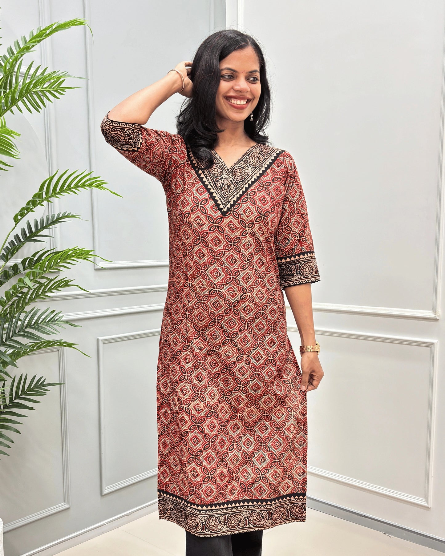 Front View of Red Women's Ajrakh Hand-block Cotton Long Kurti with red , Beige and Black ajrakh Print on V-neck ,Strip print on trim & Sleeves , standing in front of a white paneled wall with a green plant on the left.