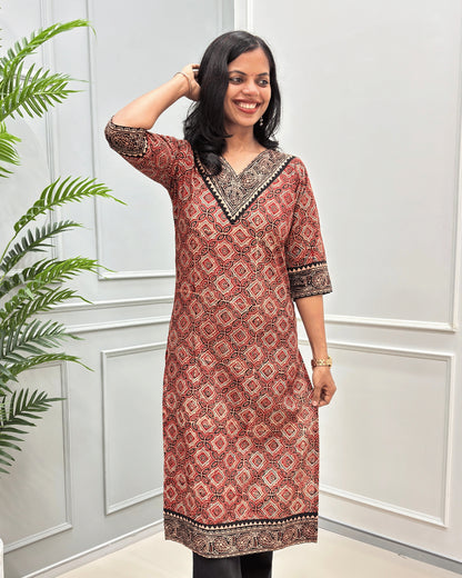 Front View of Red Women's Ajrakh Hand-block Cotton Long Kurti with red , Beige and Black ajrakh Print on V-neck ,Strip print on trim & Sleeves , standing in front of a white paneled wall with a green plant on the left.