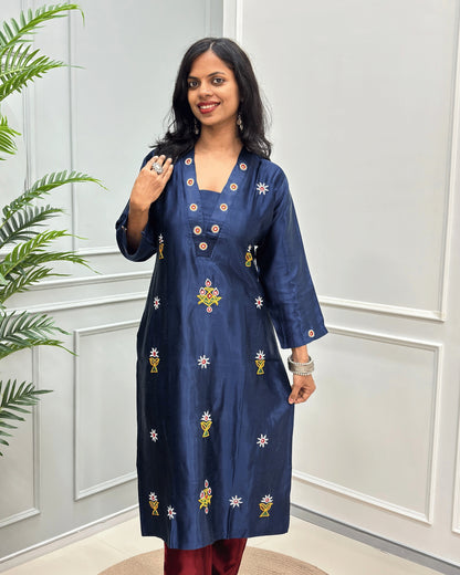 Front view of Navy Blue women's pure Mashru Silk with embroidered Ahir motifs, Hand-mirror work on front, V shaped neck, & Sleeves cuff,  standing in front of a white paneled wall with a green plant on the left.