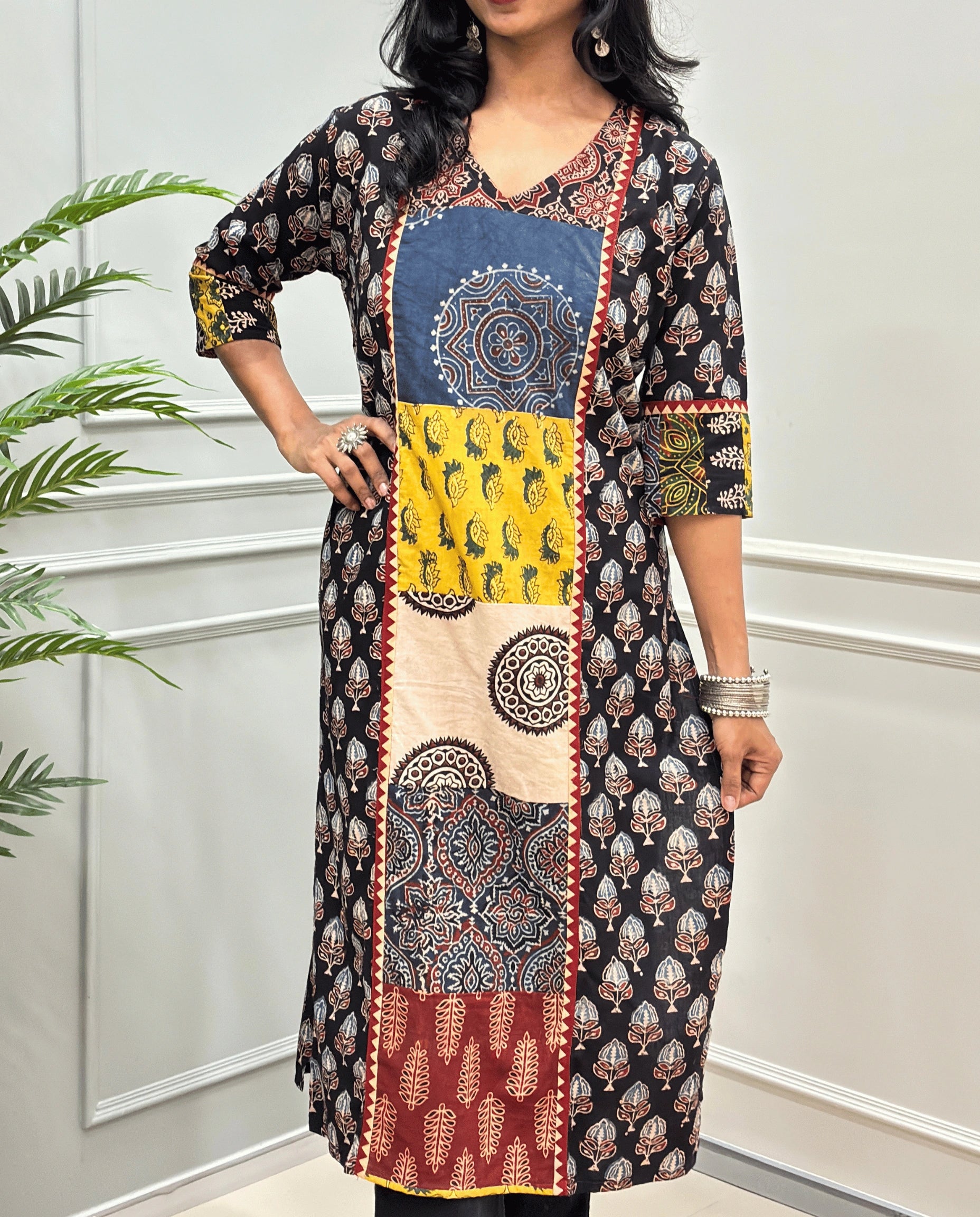 Close-up of Black Women pure cotton long kurti featured with blue, yellow, red, and black Geometric different print panels on Front & Sleeves cuffs , Ajrakh print on blue base outside the middle panel , three quarter Sleeves & V-neck standing indoors against a light paneled wall with a green plant on the left.