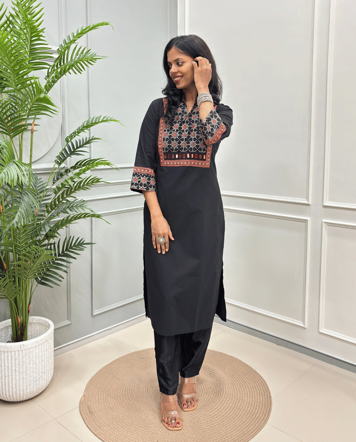 Black Women's Hand-block Ajrakh Pure Cotton Long kurti with intricate red & black geometric print on the chest and sleeve cuffs, paired with black pants , standing on a round woven mat in front of a white paneled wall with a potted plant.