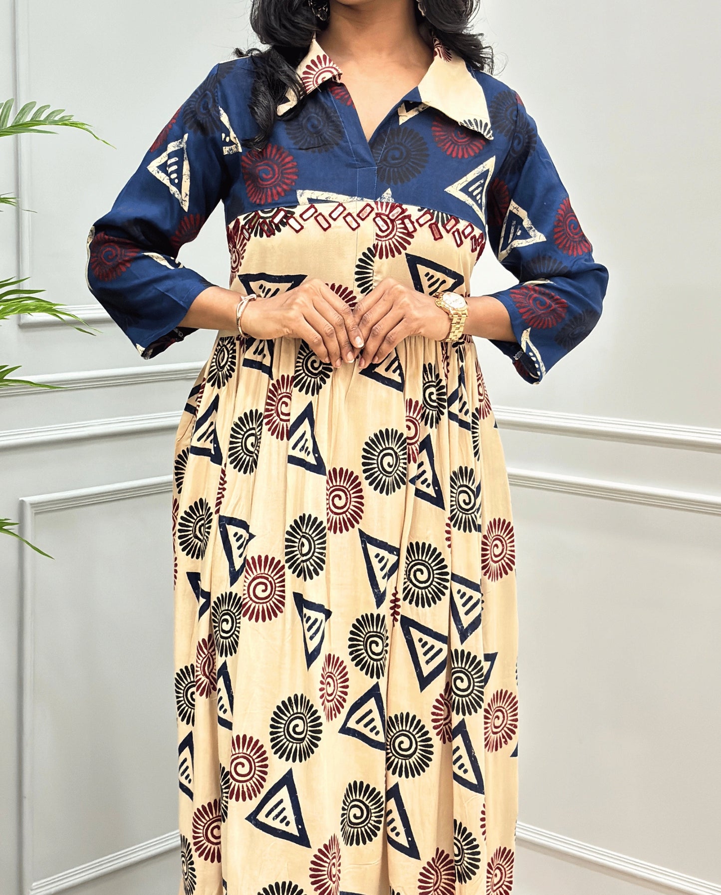 Beige Women's Ajrakh Vegetable dyed plant based Cotton Long-Kurti Featured with Three-Quarter Sleeves with a Blue neck and cuffs, and a Beige-colored collar & dress featuring a geometric pattern of triangles and spiral in Red, black, and blue, Hand-mirror work on front, standing on a round woven rug in front of a white paneled wall with a green potted plant on the left.