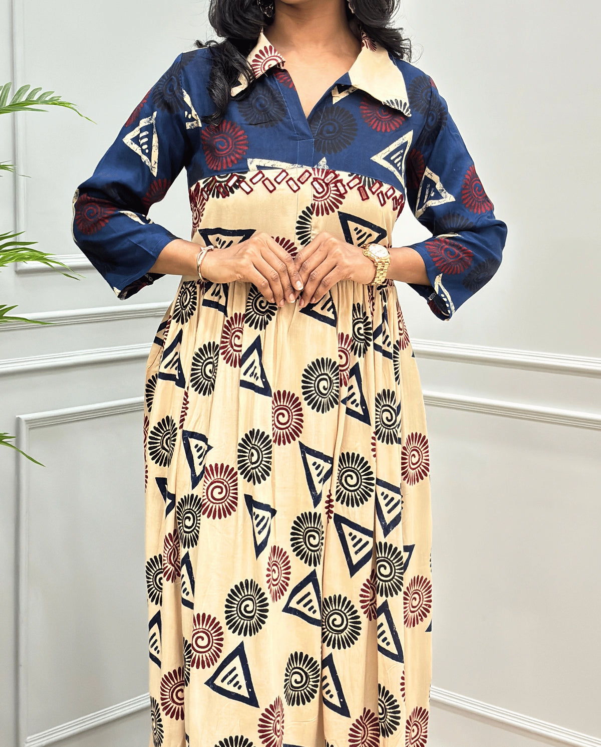 Beige Women's Ajrakh Vegetable dyed plant based Cotton Long-Kurti Featured with Three-Quarter Sleeves with a Blue neck and cuffs, and a Beige-colored collar & dress featuring a geometric pattern of triangles and spiral in Red, black, and blue, Hand-mirror work on front, standing on a round woven rug in front of a white paneled wall with a green potted plant on the left.