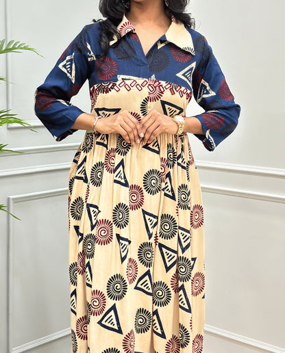 Beige Women's Ajrakh Vegetable dyed plant based Cotton Long-Kurti Featured with Three-Quarter Sleeves with a Blue neck and cuffs, and a Beige-colored collar & dress featuring a geometric pattern of triangles and spiral in Red, black, and blue, Hand-mirror work on front, standing on a round woven rug in front of a white paneled wall with a green potted plant on the left.