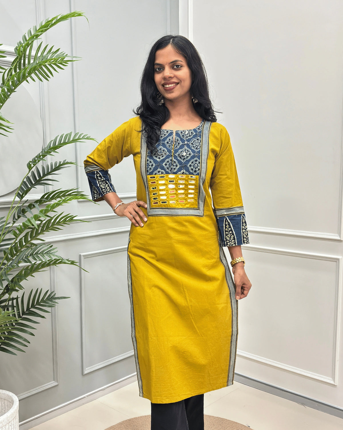 Front View of Yellow Women's Hand-block Ajrakh Pure Cotton Long Kurti with a red base and abstract Blue, Black & Beige geometric embroidery on the chest and sleeve cuffs, Hand-mirror work on Front, Ajrakh print border on Sides & Front  paired with black Flared pants, standing in a decorated room with a potted plant.