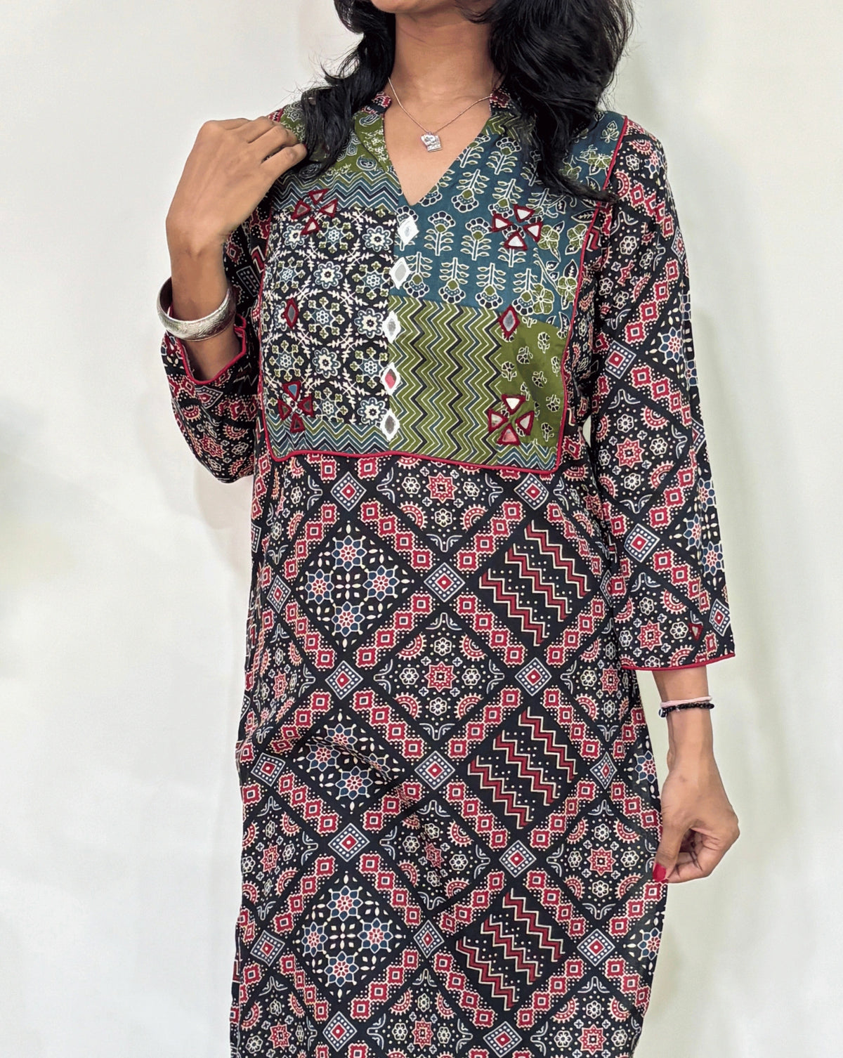 Close-up of Black Women's Ajrakh Vegetable Dyed plant based cotton long kurti Featured with full sleeves, with green, black, and red patterns, Hand-mirror work on front & Sleeves , with Mandarian V-shaped neck, paired with black flared pants, standing in a room with a white wall and a potted plant.