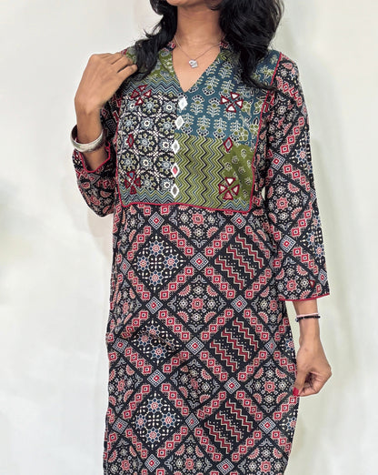 Close-up of Black Women's Ajrakh Vegetable Dyed plant based cotton long kurti Featured with full sleeves, with green, black, and red patterns, Hand-mirror work on front & Sleeves , with Mandarian V-shaped neck, paired with black flared pants, standing in a room with a white wall and a potted plant.