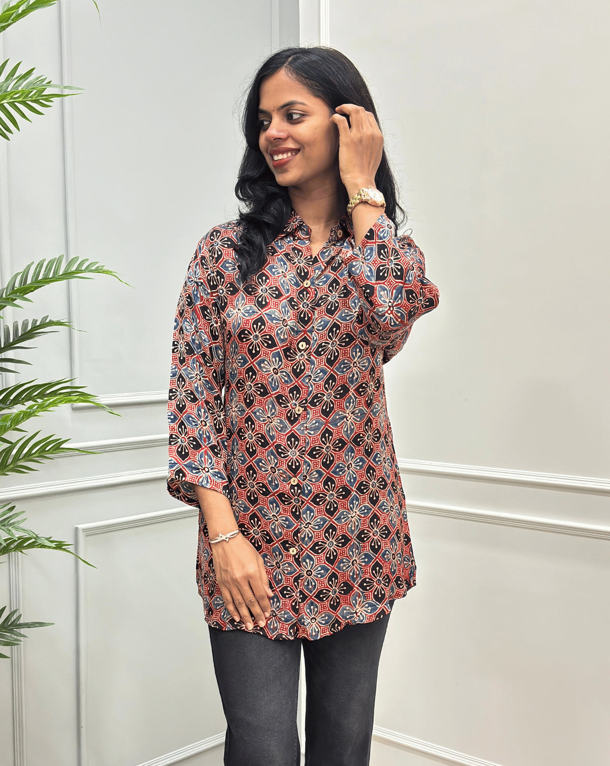 Front View of Red Women's Ajrakh Pure Modal Silk Shirt with a Floral print on blue, Black, and Red geometric pattern, black flared pants, standing on a round woven rug in front of a white paneled wall with a potted plant on the left