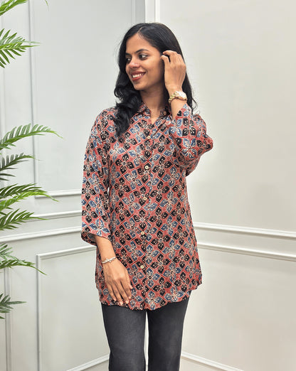 Front View of Red Women's Ajrakh Pure Modal Silk Shirt with a Floral print on blue, Black, and Red geometric pattern, black flared pants, standing on a round woven rug in front of a white paneled wall with a potted plant on the left