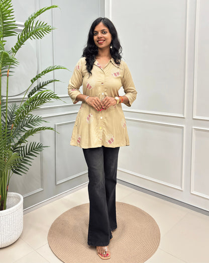 Beige Women's Ajrakh Vegetable dyed Plant based A-line cotton Short kurti with pink & white floral embroidery, paired with black flared jeans, standing on a round woven rug in a modern room with a potted plant.