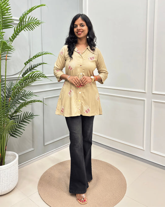Beige Women's Ajrakh Vegetable dyed Plant based A-line cotton Short kurti with pink & white floral embroidery, paired with black flared jeans, standing on a round woven rug in a modern room with a potted plant.