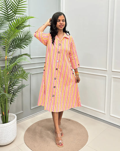 Multi Women's Ajrakh A-line Pure Cotton Long Kurti with an Ajrakh Strip Print on abstract of Pink, White, Yellow, Orange,  standing on a round woven rug in front of a light gray paneled wall with a potted plant.