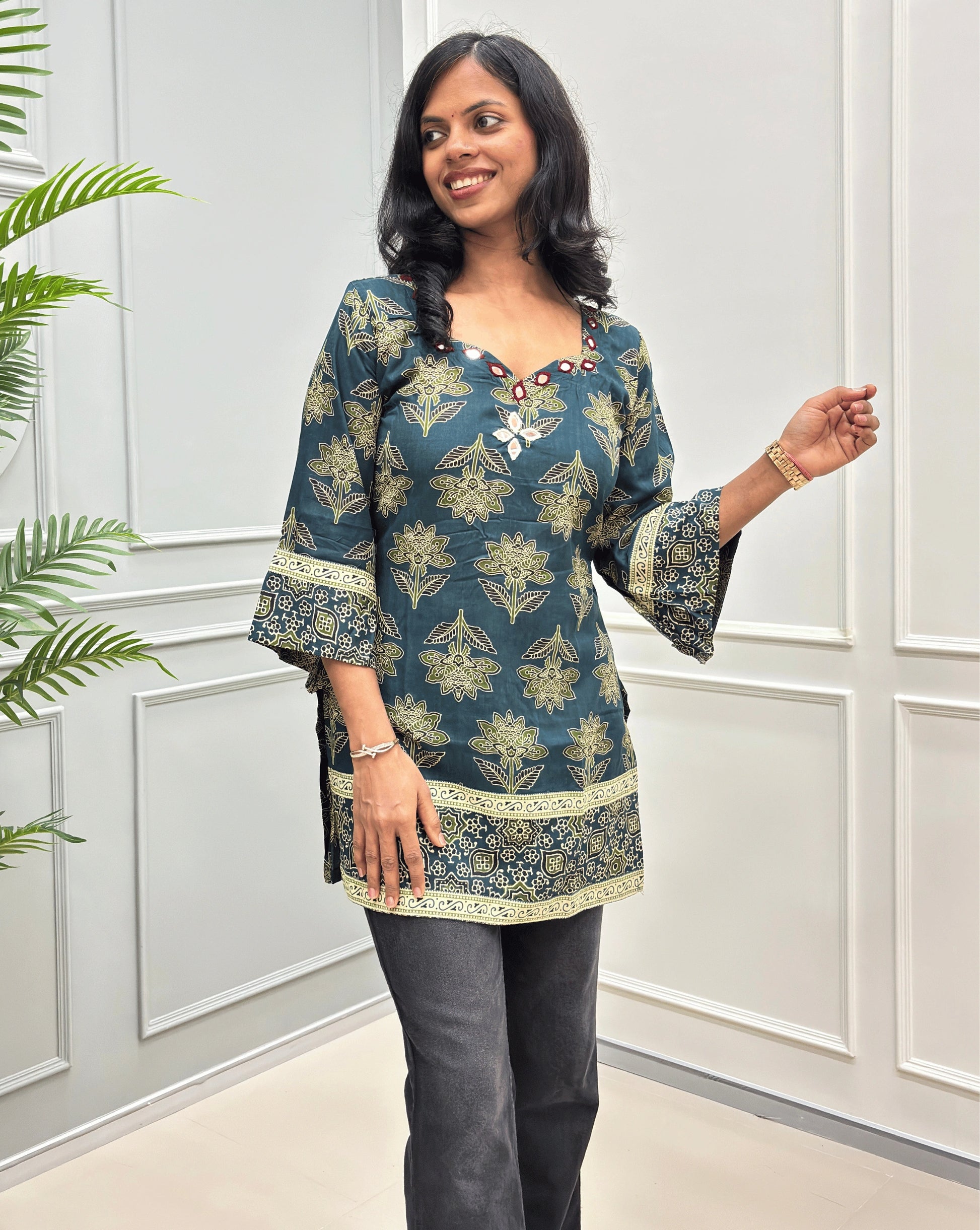 A woman modeling a teal blue and beige printed kurta with wide bell sleeves, paired with dark gray pants, standing in front of a white paneled wall.