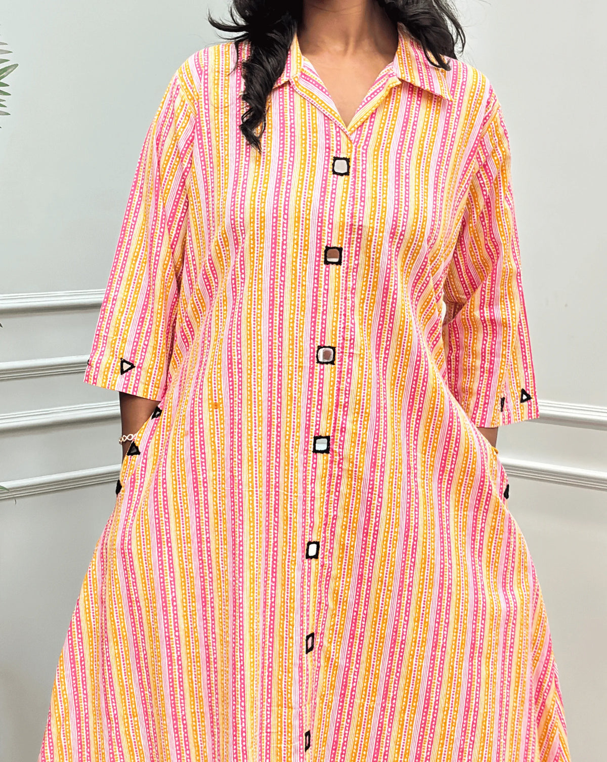 Close-up View of Multi Women's Ajrakh A-line Pure Cotton Long Kurti Featured with Collar V neck & 3/4th Quarter Sleeves with an Ajrakh Strip Print on abstract of Pink, White, Yellow, Orange, Real hand-mirror work on front & Sleeves, Along with 2 side pockets, standing on a round woven rug in front of a light gray paneled wall with a potted plant.